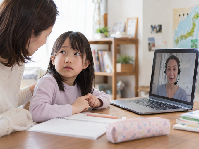 パソコンの前で言葉に詰まり隣の親を不安そうに見つめる小学生のオンライン英会話風景
